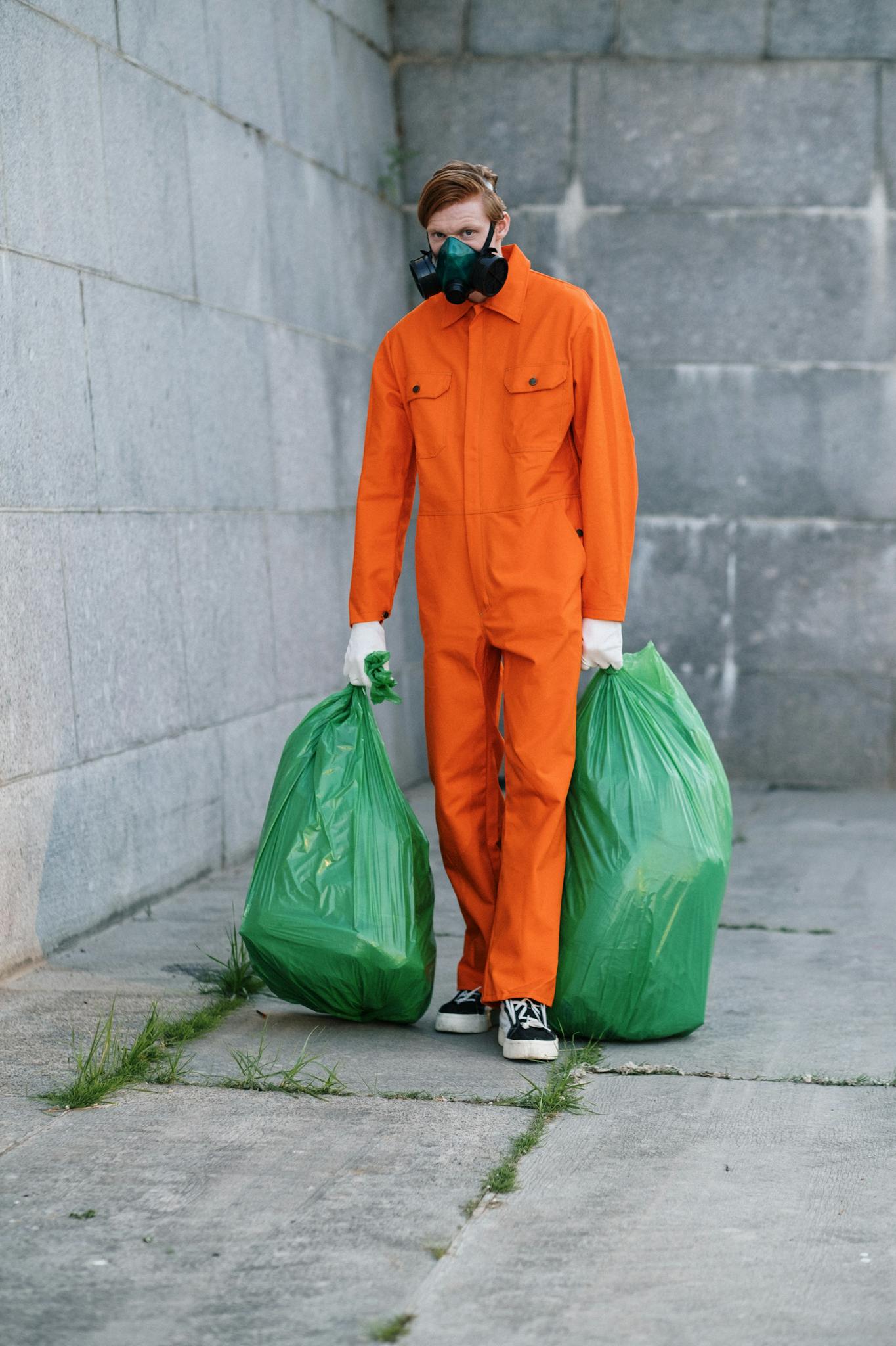 A man in an orange jumpsuit and mask carrying garbage bags against a concrete wall.